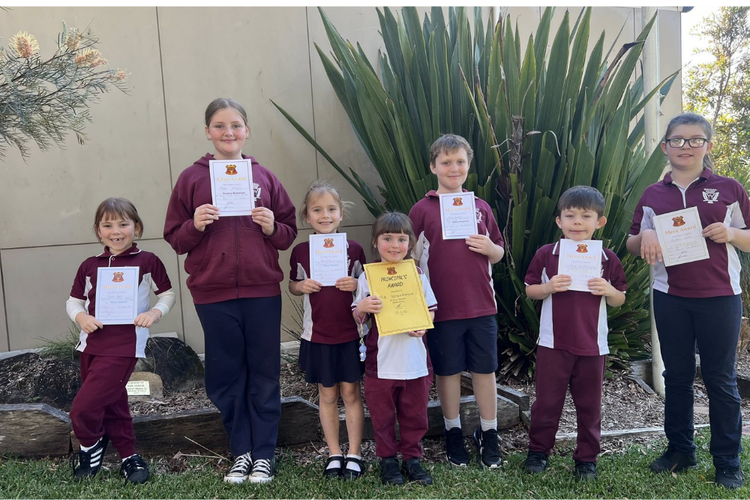 Seven students of mixed ages standing in uniform holding awards received at assembly.