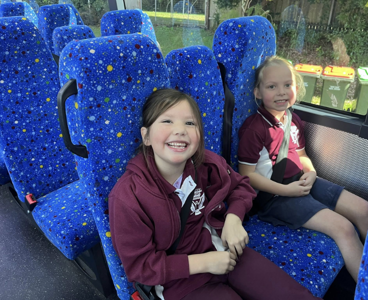 Two smiling young female students sitting on blue bus seats going on an excursion.