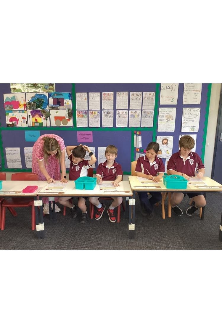 A row of four young students at desks with a teacher helping one with his work