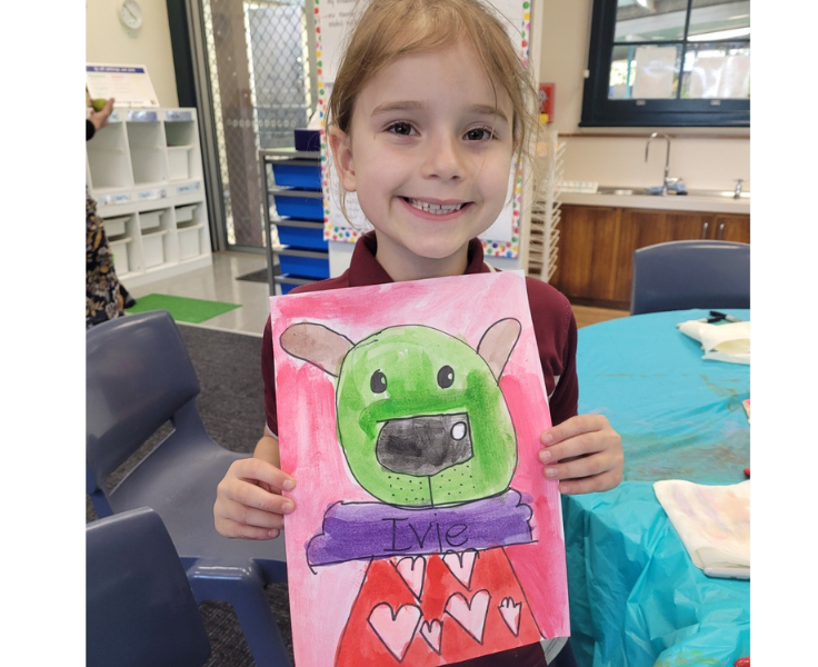 A year 1 female student holding a colourful artwork of a green monster sitting on a purple log.