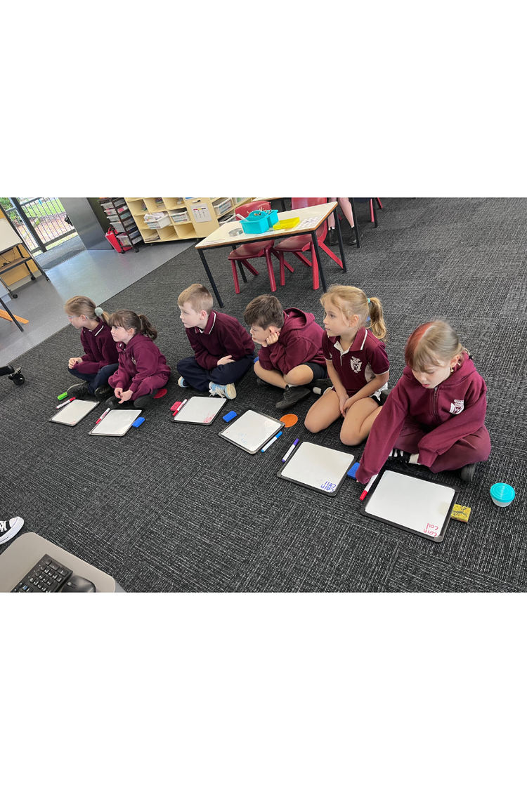 Students in K-2 on floor with small whiteboards listening to teacher