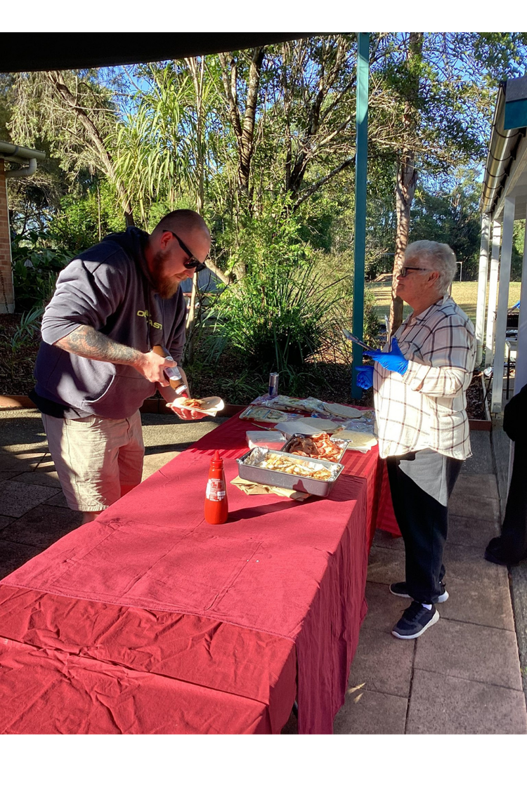 Two parent/carers at a long table helping themselves to a sausage sandwich for Open Day Education Week celebrations.